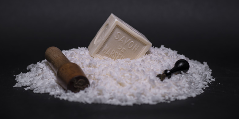 Cube de Savon de Marseille de 600g sur copeaux et entouré de tampons à savon sur fond noir