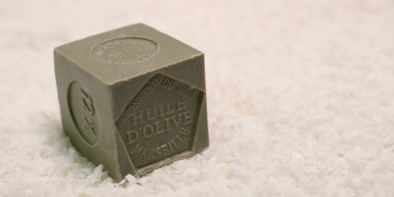 Cube de savon de Marseille à l'huile d'olive, dont il prend la couleur olive verte, produit par la Savonnerie du Midi et posé sur tapis de copeaux de savon